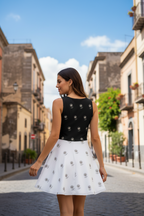 Woman in a black top and white skirt standing on a sunlit street with buildings and plants in the background.