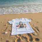 White t-shirt with a printed design on a sandy beach with ocean waves.